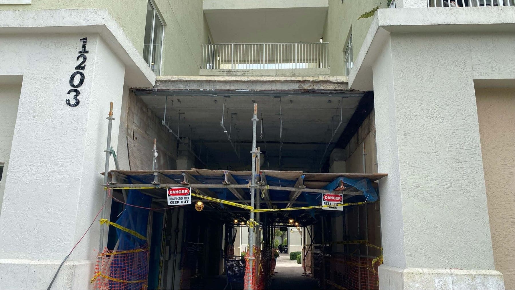 Under a balcony at the Abacoa building, the stucco soffit stripped to its framing during waterproofing repairs, with shoring and danger tape across the walkway below