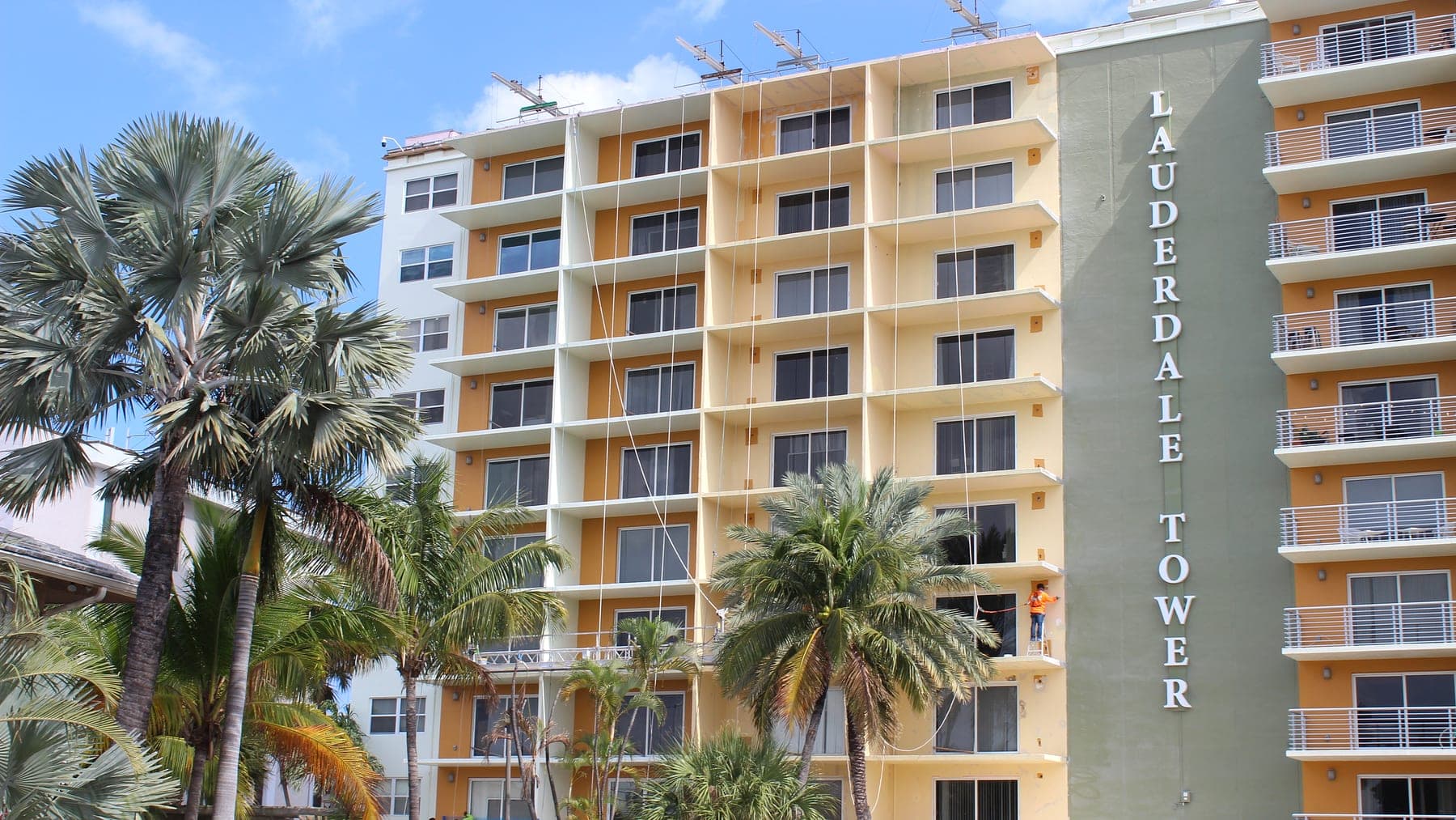 Lauderdale Tower mid-repaint, with a painter on a swing stage beside the building's vertical lettering